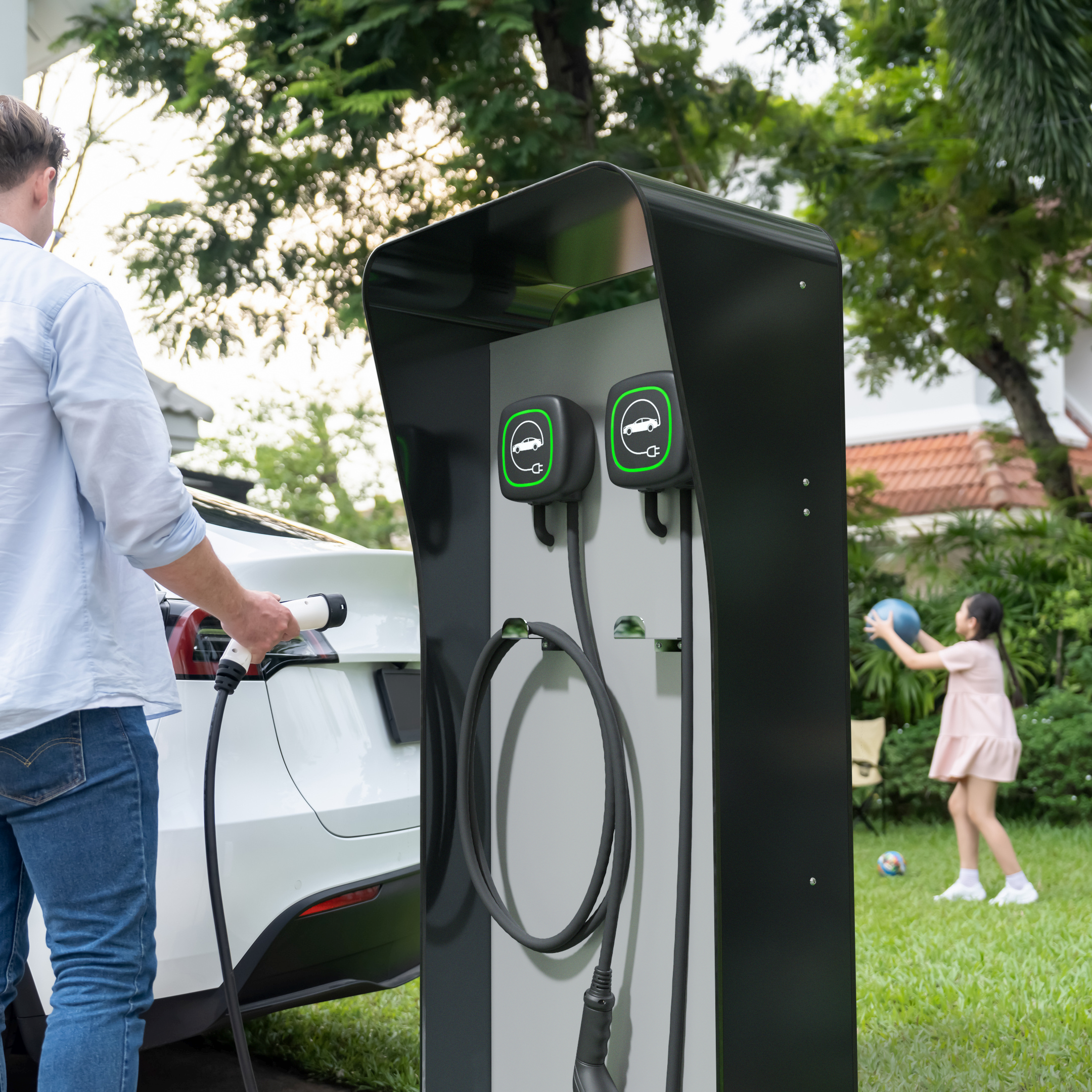 Mann lädt ein Elektroauto mit einer freistehenden Ladestation mit zwei Wallbox-Halterungen. Die Säule steht im Garten eines Wohnhauses.